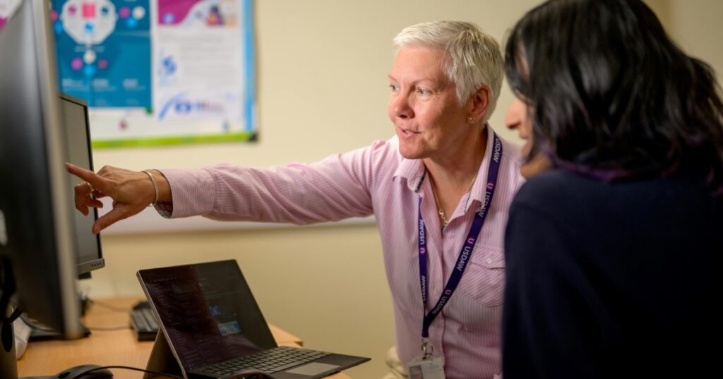 A tutor helps a learner with digital skills training. Both are sat down at a desk with computer in front of them.