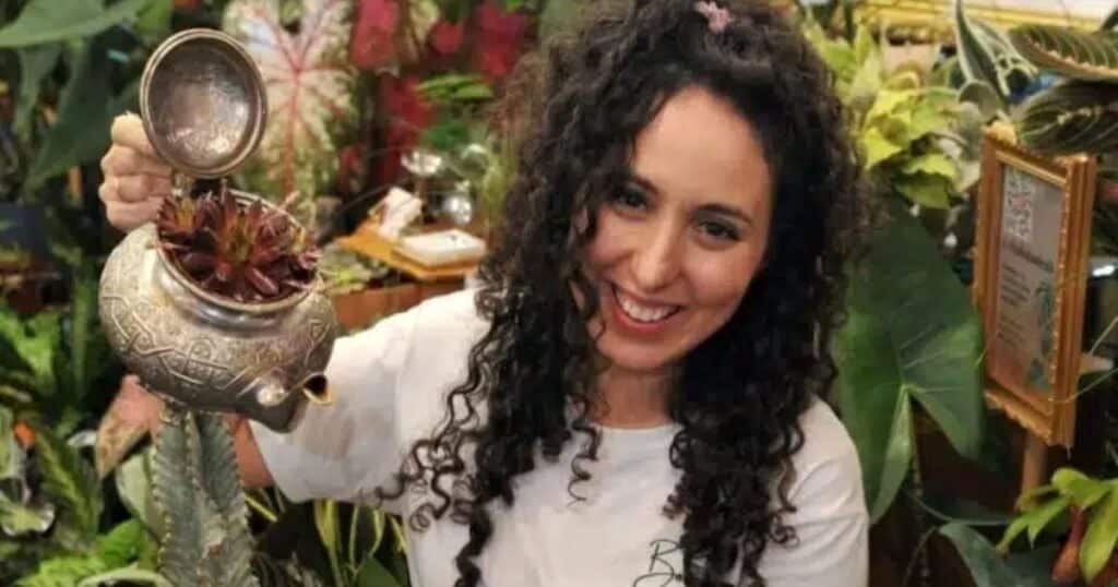 Headshot of a woman with dark hair holding a silver teapot in her hand. She's surrounded by flowers and botanicals.