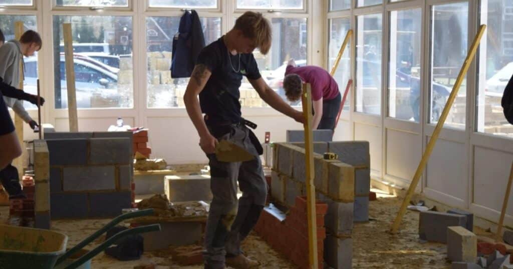 A construction worker stands looking at a newly-built wall. He holds a trowel in one hand and a spirit level in the other.